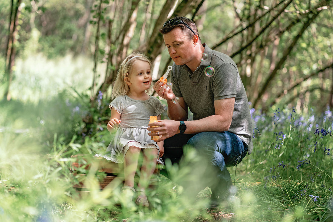 Girl sat on a box surrounded by bluebells blowing bubbles next to her dad